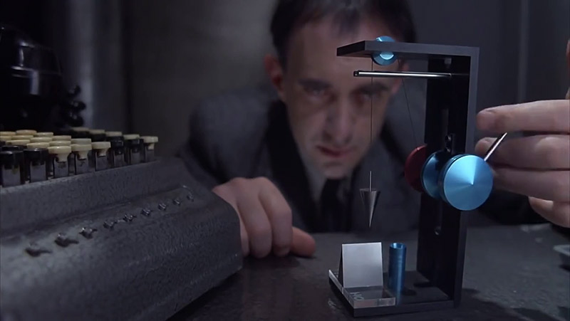 Sam Lowry leans over a desk, turning the crank of a small decision-making machine that drops a suspended weight toward “Yes” or “No.”